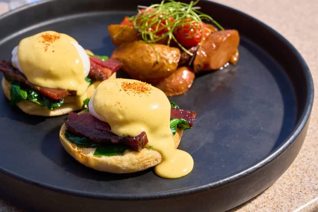 S3 Benedict at S3 Restaurant in Fort Lauderdale, served with poached eggs, maple-glazed bacon, sautéed spinach, and miso hollandaise over English muffins with crispy potatoes